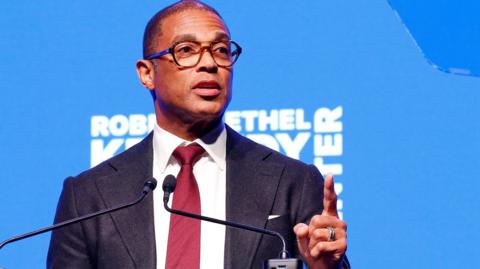 Don Lemon wearing a dark coloured suit, white shirt and red tie. He is standing in front of a bright blue background. 