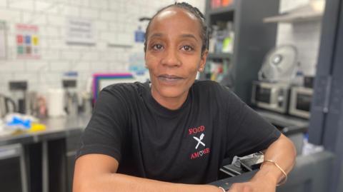 Michelle Witter. She is standing with her arms resting on a surface. She is wearing a black T-shirt with a white and red Food Amore logo on the chest. She is looking directly at the camera and smiling 