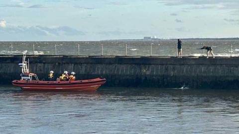 Three lifeboat crew members in a lifeboat close to a harbour wall. Two people can be seen on the harbour wall calling out to a dog that has fallen into the water.