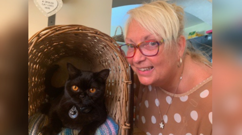 The image shows a woman smiling while posing with her black cat, who has amber eyes and is sitting in a basket.