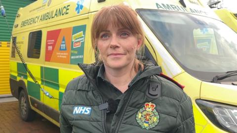 Chrissy Ames is stood in front of an ambulance. She is wearing a dark green puffa jacket with a blue NHS badge sewn onto one side and the NHS Ambulance Service crest badge on the other.