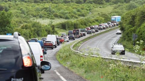 Long queues of cars on one carriageway. The other carriageway closed with surrounding hills. 