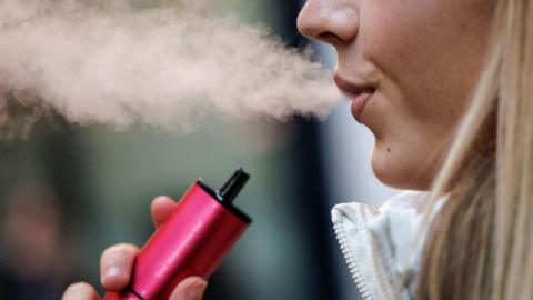 A woman with blonde hair exhaling vape smoke while holding a pink vape