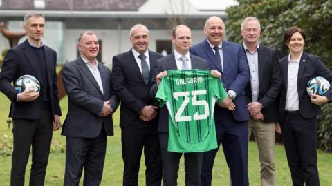 Gordon Lyons who is balding with reddish brown hair at the sides and who is wearing a dark suit holds a Northern Ireland football top with Galgorm 25 on the back of it. Standing beside him are five men and one woman, including Northern Ireland football manager Michael O'Neill and form international Aaron Hughes.