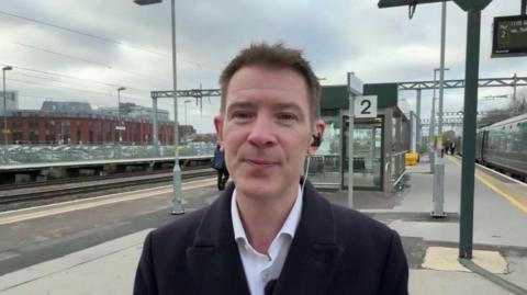 James Davis wearing a dark navy blue overcoat with a white shirt that is unbuttoned at the collar. He is clean shaven with short brown hair. He is also stood at a railway station with trains and passengers moving behind him.