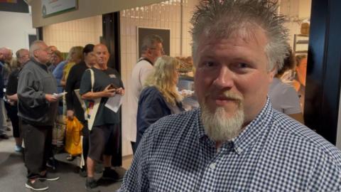A man with a beard, wearing a blue and white checked shirt, is standing in front of a queue in a market. 