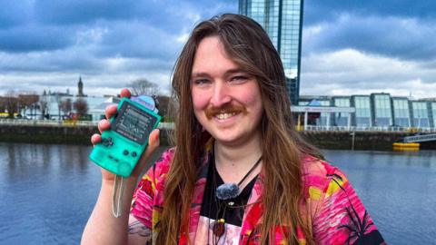 A man in a colourful pink shirt, holding up a Game Boy and smiling at the camera