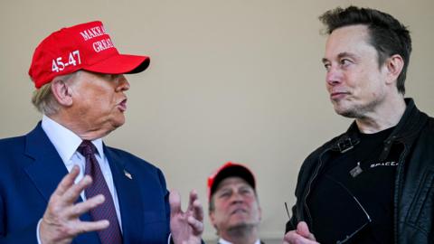 Donald Trump, wearing a red baseball cap with the numbers 45-47 on the side and a navy suit, in side profile as he speaks to Elon Musk, wearing a black leather jacket and black T-shirt