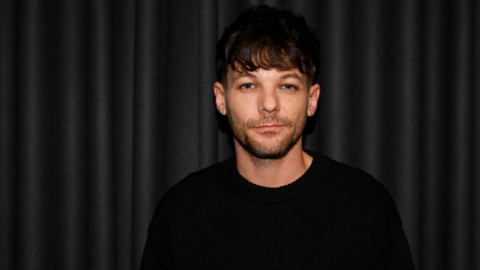 A man wearing a dark sweater stands in front of a black curtain.