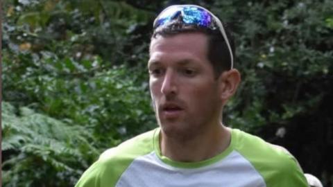 A man, with short brown hair, wearing sunglasses. He is wearing a white top with green shoulders. Behind him is green tree branches with leaves. 