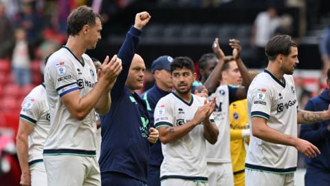 Millwall players in white kit clap whilst head coach Alex Neil raises his right fist in the air with a determined look on his face