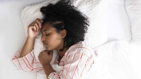 A woman sleeping on her side with her arms near her face in a bed with white bedsheets.