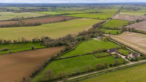 An aerial view of green land. Acres of land can be seen lined with trees and there is a road and a couple of houses also visible.