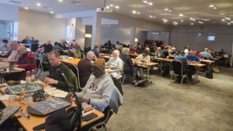 A room of people sitting at long tables, with their heads down, looking at big Scrabble boards 