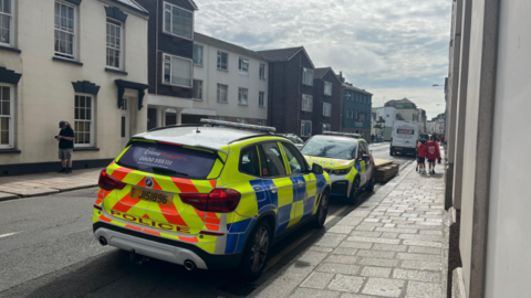 A row of buildings on a main road, two police cars parked next to the pavement.

