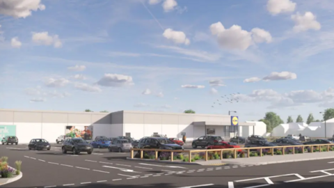 The artist impression of the new store shows a large white and grey building with a large car park in front f it with some cars parked on it. A low wooden rail surround the parking area and entrance. The Lidl sign can be seen outside the store.