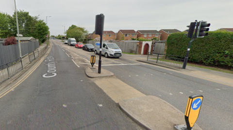 A pedestrian crossing on County Road North in Hull, there are traffic lights and queuing cars.