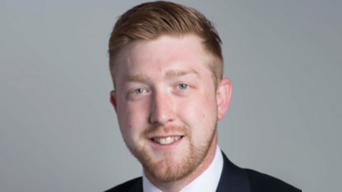 Corporate headshot of a man with reddish-brown hair and a trimmed beard and moustache smiling at the camera. A white short collar and black jacket are just visible at the bottom of the shot.