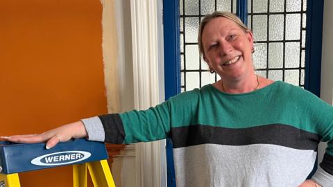 Exec Director of The Garage Trust Carrie Mansfield stands in a doorway leaning on a yellow step ladder. She is wearing a green and grey top. Her blonde hair is tied back.