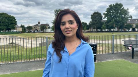 A woman with shoulder-length brown hair, wearing a light blue blouse. She is stood in a park, and behind her is a skating area, grass and trees.
