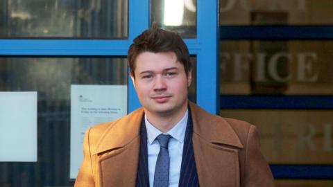A man with brown hair, wearing a brown overcoat and a blue suit. He looks at the camera while standing outside a blue door.