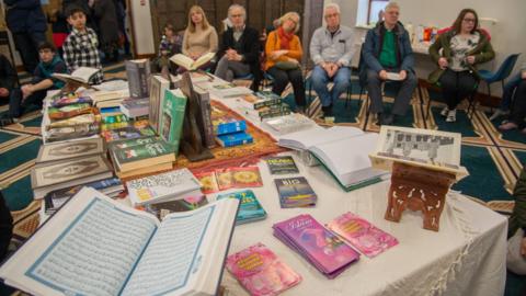 A picture of an open day from a previous year with a Quran and other Islamic books with people sitting on chairs in the background 