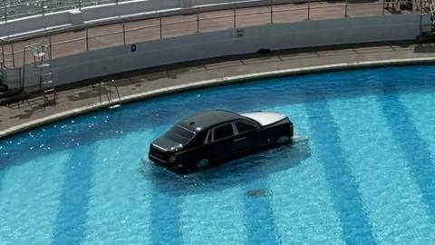 A Rolls Royce car on a submerged plinth in Tinside Lido in Plymouth on a sunny day. A fountain in the middle of the pool is spouting out water. The car is mostly black with a silver bonnet. A film crew is gathered on the outside edge of the pool.