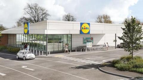 A CGI artist impression of the Lidl store which is a large, white building with a partial glass front on the left side. Two blue and yellow Lidl logo sit on the roof of the roof of the store, one facing the car park on the right, and the other facing a main road on the left. There are cars in the car park and two driving down the road.