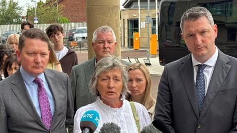 Shows a woman with grey hair and a white top standing in front of microphones. She if flanked by two men in suits and other people are in the background.