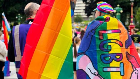 Two men draped in rainbow flags, one wearing a rainbow hat, stand with their backs to the camera