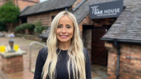 A woman with long blonde hair and a black top stands outside a building smiling at the camera