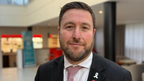 Pete Marland with short dark hair and a beard, wearing a white shirt, red tie, black jacket. He is standing in front of a yellow and blue wall.