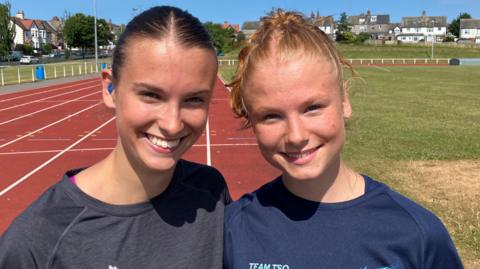 Honor and Getta smiling into the camera while on a red running track. Honor is on the left and has brown hair which is tied back. She has a blue hearing aid in her ear and is wearing a grey t-shirt. Next to her, Gretta has strawberry blonde hair which is also tied back and is wearing a blue t-shirt. There is a patch of grass in the middle of the track and houses in the background.