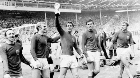 Geoff Hurst, who is wearing a long-sleeved football shirt and shorts, is holding the World Cup Trophy while standing next to a number of other players on the pitch. The stands are filled with fans in the background.