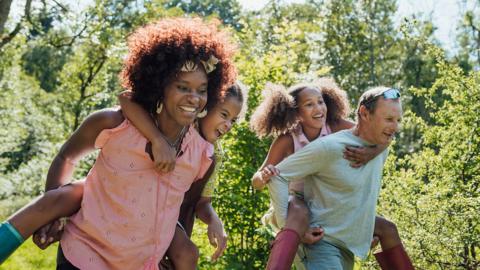 A woman and a man smiling while each caries a laughing child on their back, with green foliage visible behind
