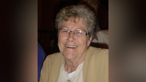 Beryl Barrett, with short hair and glasses, smiles at the camera and is wearing a cream cardigan.
