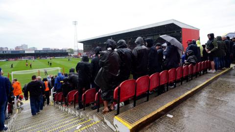 A view of the County Ground football stadium in Swindon from the stand looking down to the pitch. People are standing up watching a game. It is raining and spectators are dressed in black rain coats.