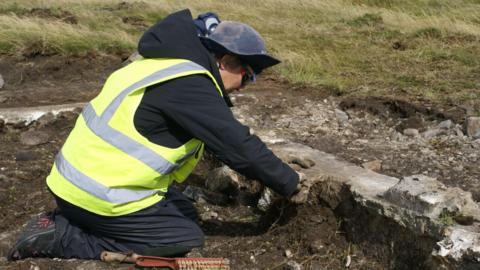 A man wearing a hi-vis vest helps excavate the foundations of a Nissen hut from the Second World War