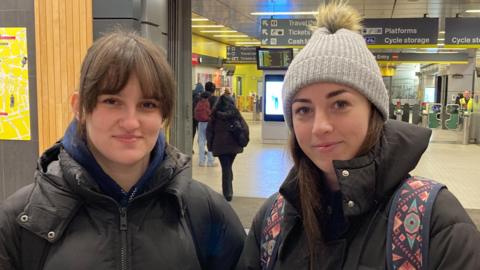 Orlas and Ellie are standing inside Liverpool Central station. Orla has brown hair and is wearing a black winter coat. Ellie has long brown hair and is wearing a grey woolen hat and a black winter coat and a pink and blue patterned rucksack.