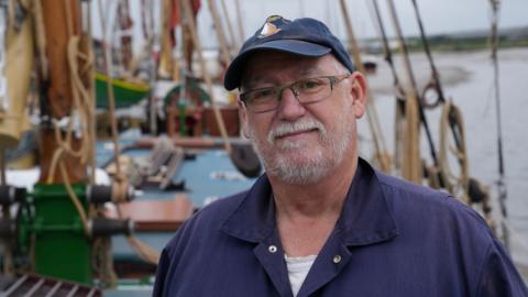 Tim Kenney is dressed in a navy sailor's jacket, wearing a navy baseball cap, and standing on the deck of Pudge the Thames sailing barge