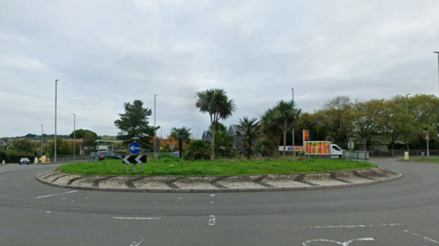 The roundabout on the A390 on a cloudy day. The islands has palm trees and grass as well as road traffic signage. There are two cars and a van on the opposite side of it and the lanes in the foreground are empty.