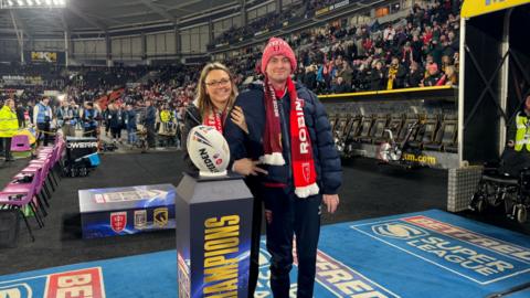 Craig Eskrett and his wife standing beside a display plinth holding a rugby ball inside a large, crowded stadium. They are both wearing warm clothing and team scarves, with spectators filling the stands behind them.