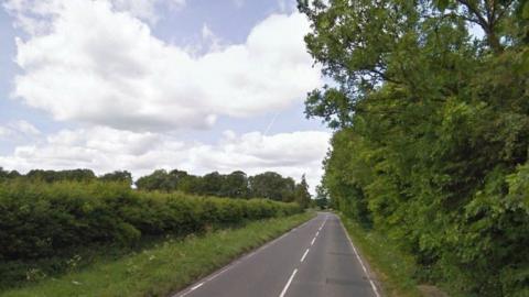 An image of a country road, the A413, which is surrounded by green bushes on either side. There are trees in the distance.