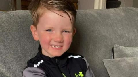 A blond boy sits on a grey sofa. He has blonde hair and blue eyes, and he is wearing a black and grey sports top. He is smiling at the camera.