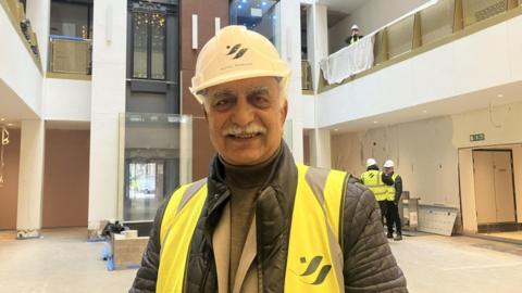 Mahboob Hussain, wearing a white hard hat, and a high visibility jacket. He is standing in one of the restaurant rooms smiling.