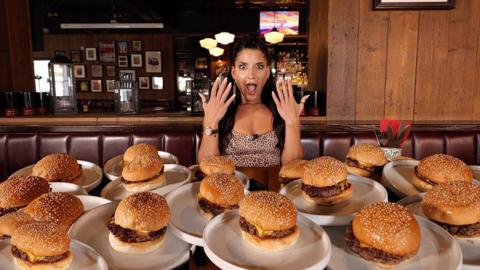 A woman sits in a restaurant with several plates with burgers on the table in front of her. She has straight dark hair and she is hold her hands up with a mock shocked expression.