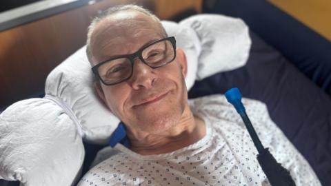 A man in a hospital gown smiling while lying on a bed