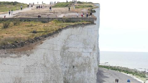 A white chalk cliff, with people stood close to the edge taking photos. Below, there is a walkway with people walking on it, and the sea in the background.
