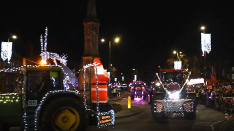 Illuminated tractor with Santa statue on the back in a convoy passinf Banbury Cross at night