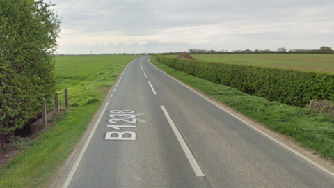 Google shot of a minor road. It has white road markings and is flanked by fields on either side. There is a ditch and a hedge to the right of the picture. 